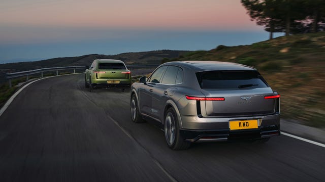 rear three-quarter view. Two luxury SUVs driving on winding mountain road at dusk with hills beyond.