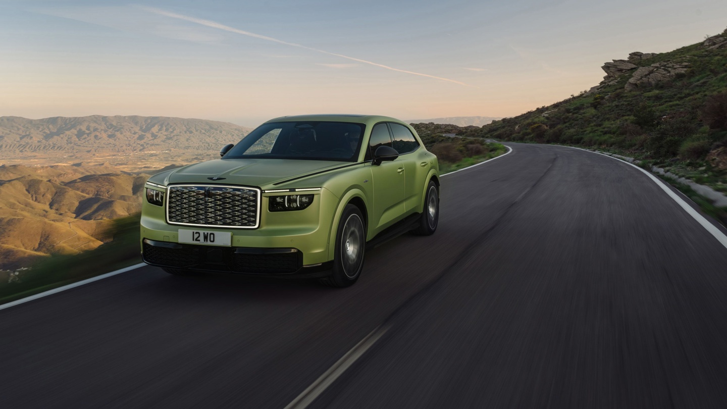 A green SUV driving on a winding road through mountainous terrain at sunrise.